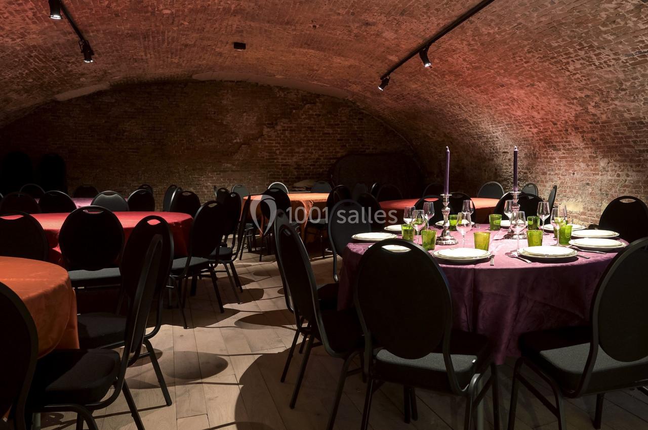 Salle voûtée en briques avec tables rondes dressées, nappes colorées et chaises noires, éclairée par des spots.