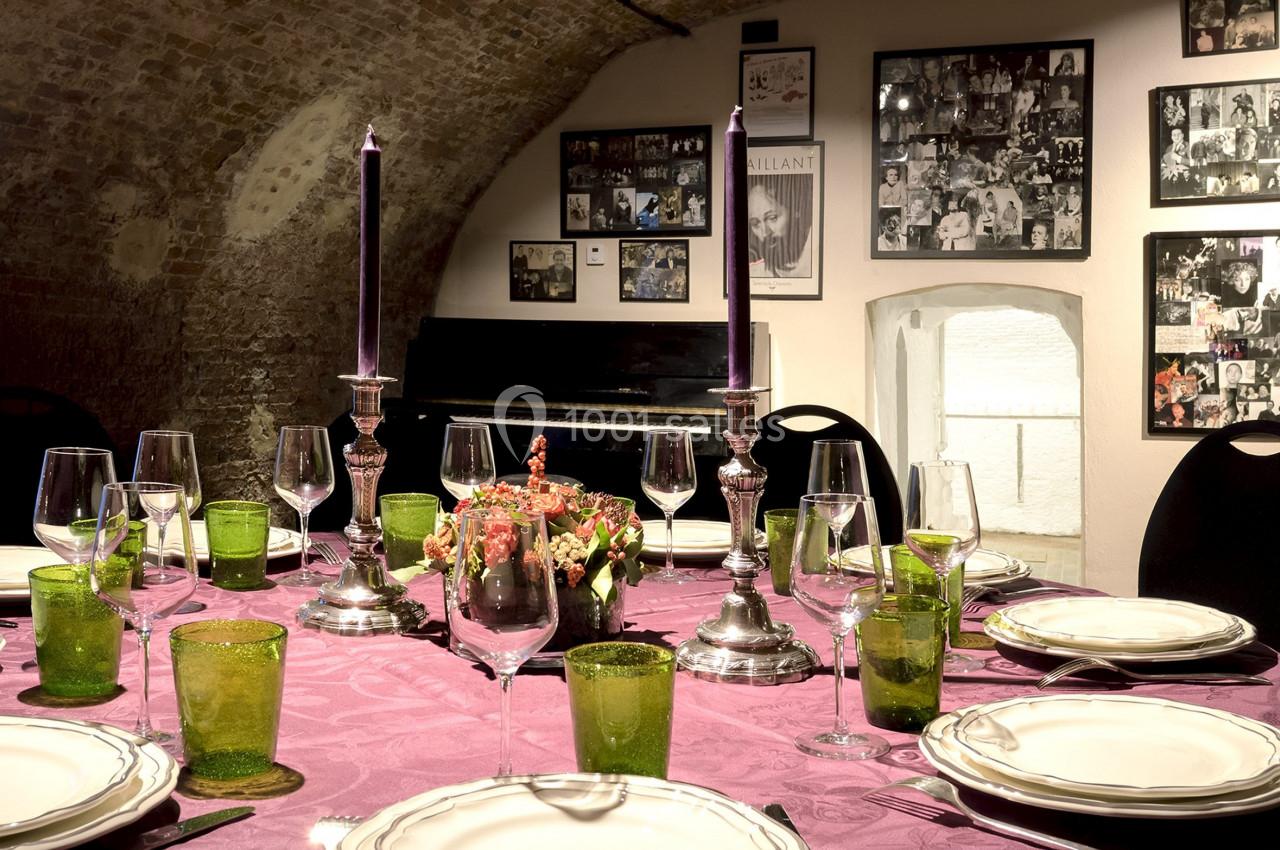 Table dressée avec nappe violette, vaisselle élégante, bougies allumées, dans une salle voûtée décorée de photos.