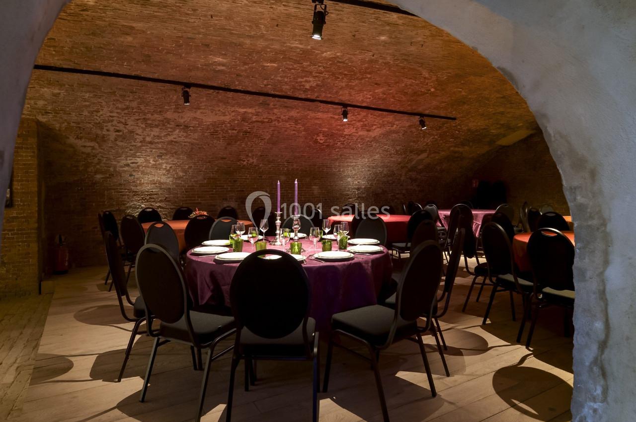 Salle voûtée avec des tables rondes dressées pour un repas, éclairée par des bougies et des lumières tamisées.