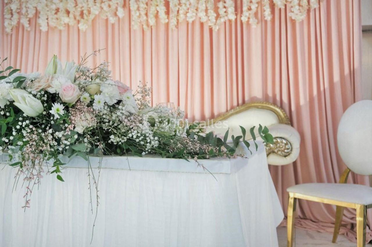Arrangement floral blanc et rose sur une table drapée, avec rideaux roses et fauteuil doré en arrière-plan.