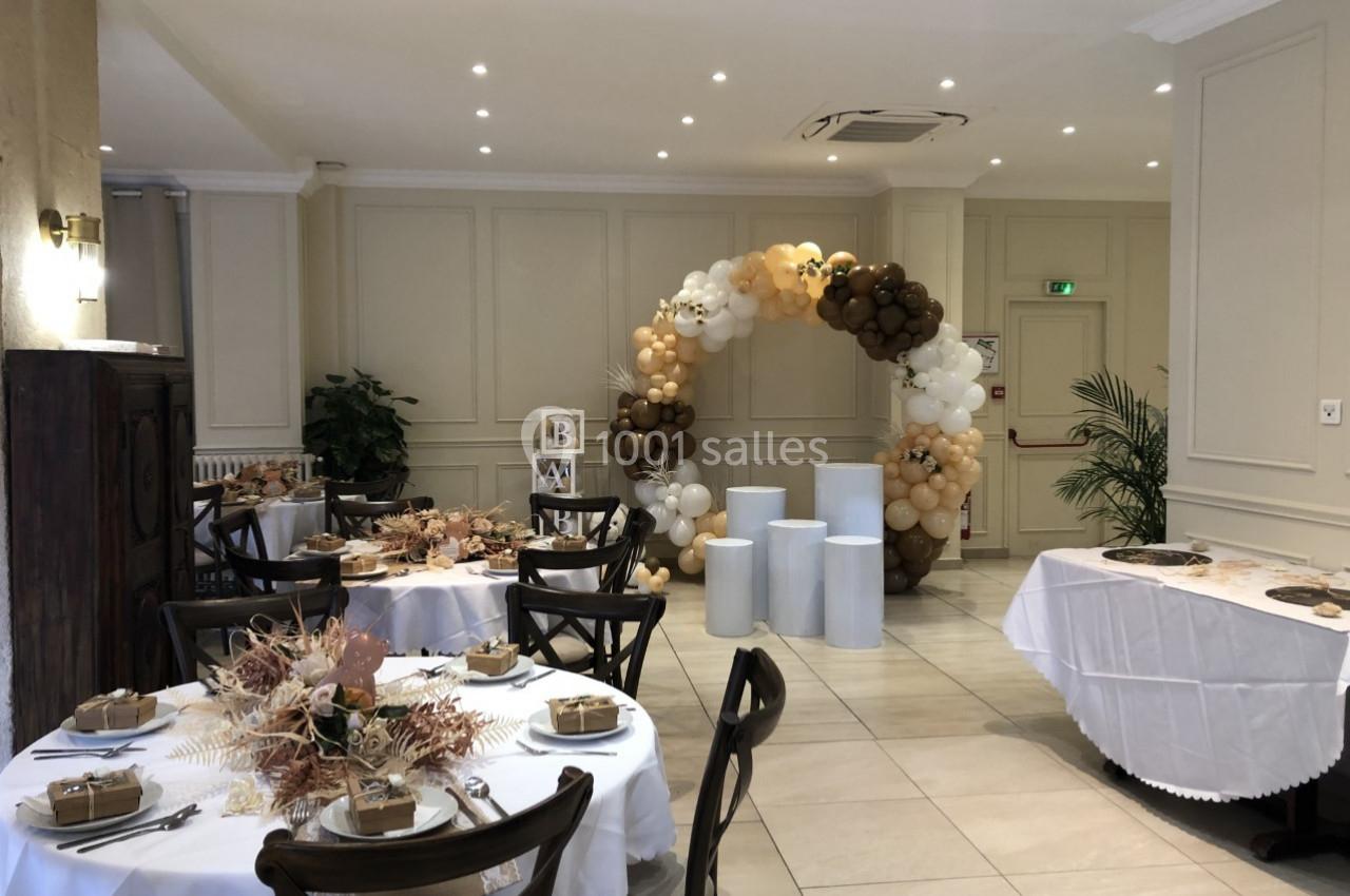 Salle décorée pour un événement avec tables dressées, arche de ballons beige et marron, et colonnes blanches.