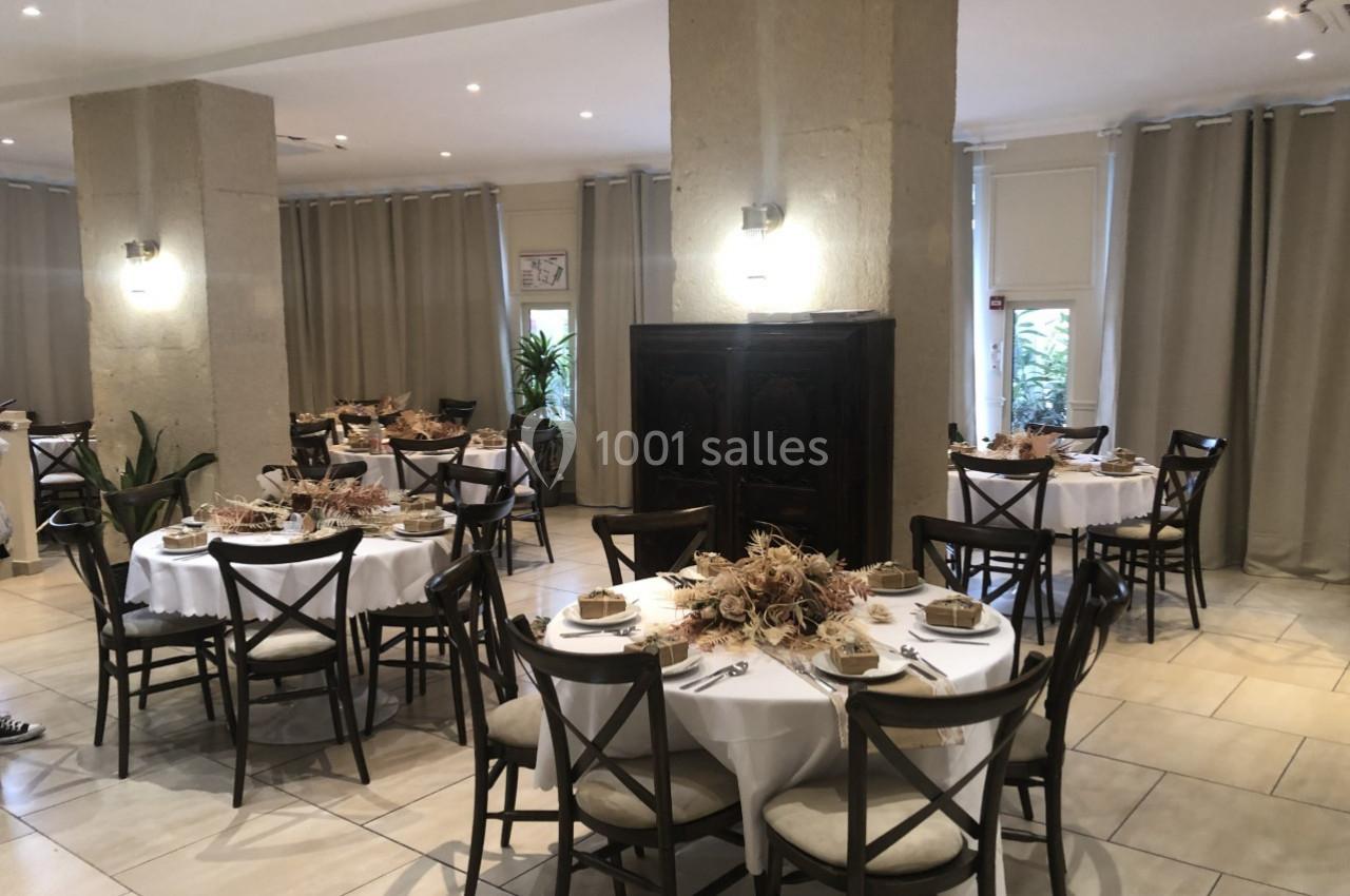 Salle de réception avec tables rondes dressées, décorées de centres de table en fleurs séchées, éclairée par des appliques…