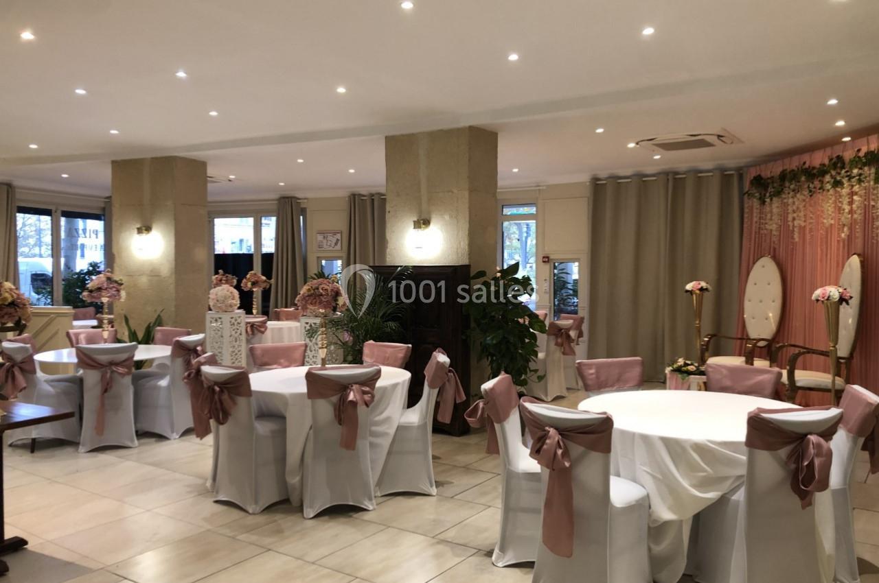 Salle de réception décorée avec des tables rondes, nappes blanches, chaises avec nœuds roses et rideaux beiges.