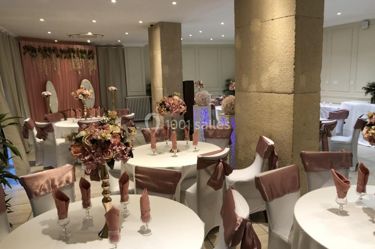 Salle de réception décorée avec des tables rondes, nappes blanches, serviettes roses et compositions florales élégantes.