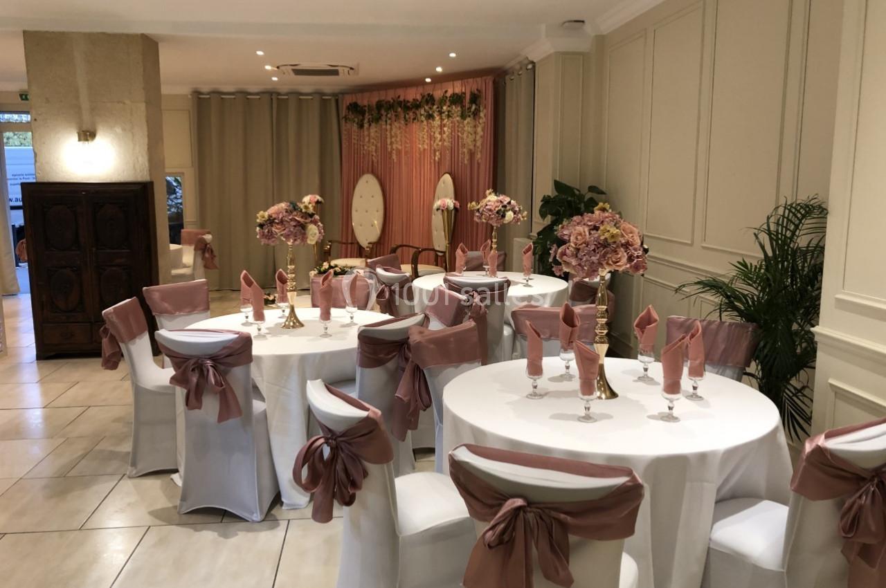 Salle de réception décorée avec des tables rondes, nappes blanches, chaises ornées de nœuds roses et compositions florales.