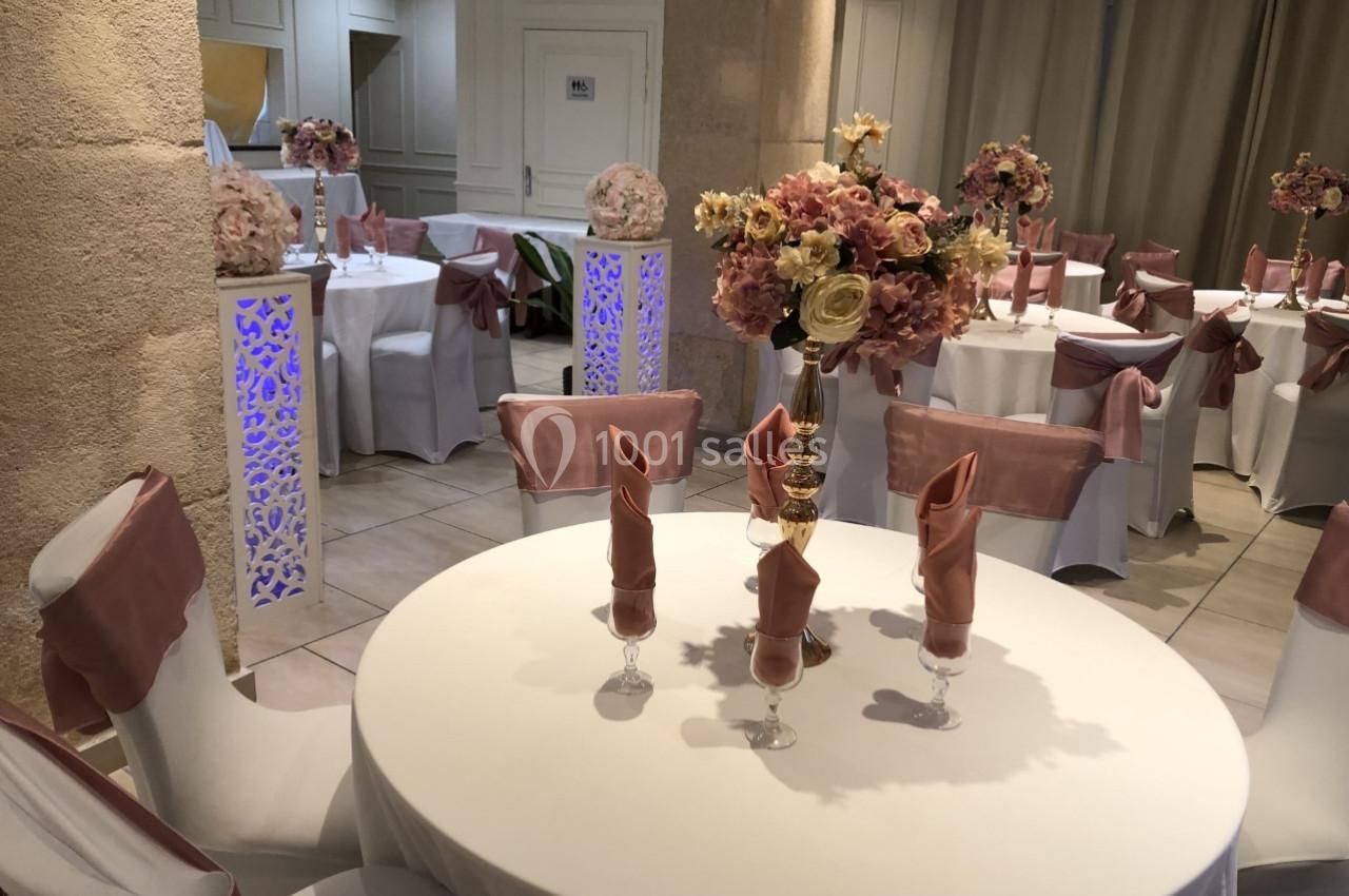 Salle de réception décorée avec des tables rondes, nappes blanches, chaises ornées de nœuds roses et centres de table…