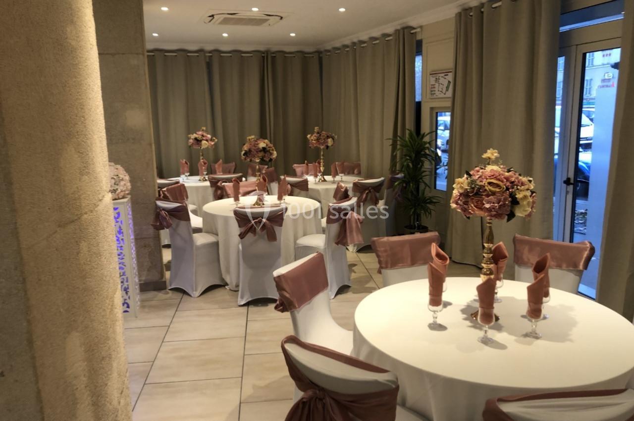 Salle décorée pour un événement avec tables rondes, nappes blanches, chaises ornées de nœuds roses et bouquets floraux.
