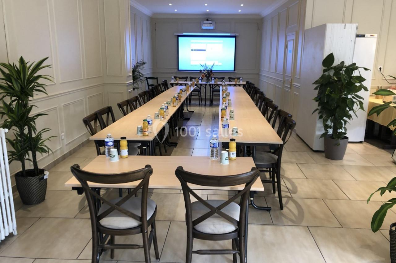Salle de réunion lumineuse avec tables disposées en U, chaises en bois, écran de projection et plantes décoratives.