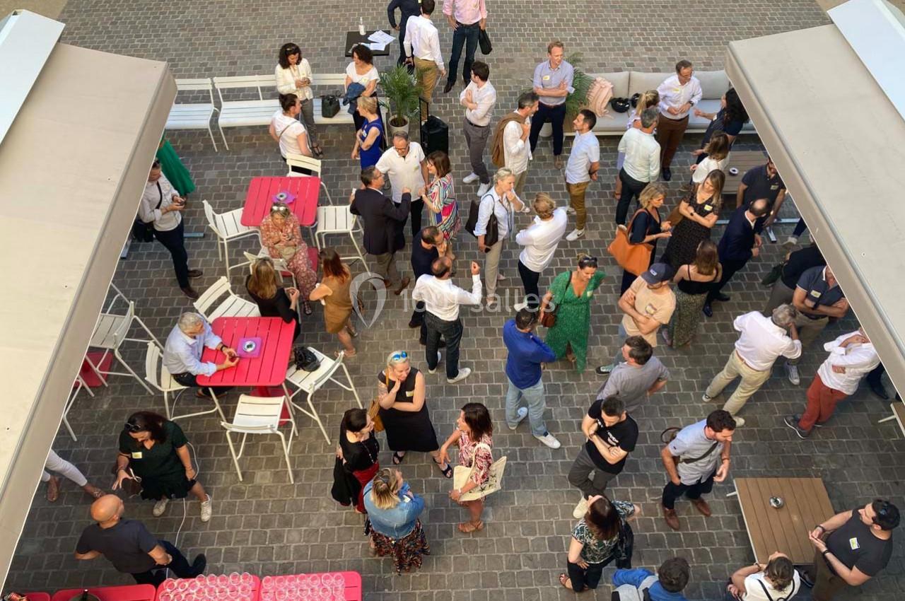 Vue aérienne d'un rassemblement en extérieur, avec des personnes discutant autour de tables sur une terrasse pavée.