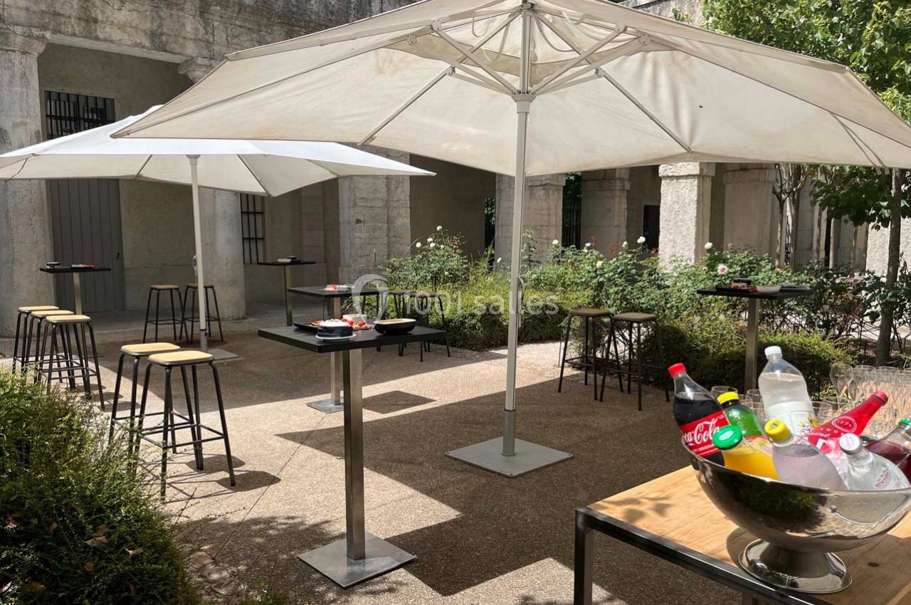 Terrasse extérieure avec tables hautes, chaises, parasols et boissons fraîches dans un seau rempli de glace.