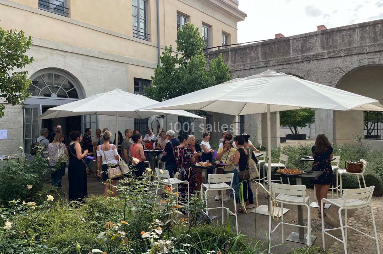 Groupe de personnes rassemblées dans une cour extérieure, discutant autour de tables sous des parasols blancs.