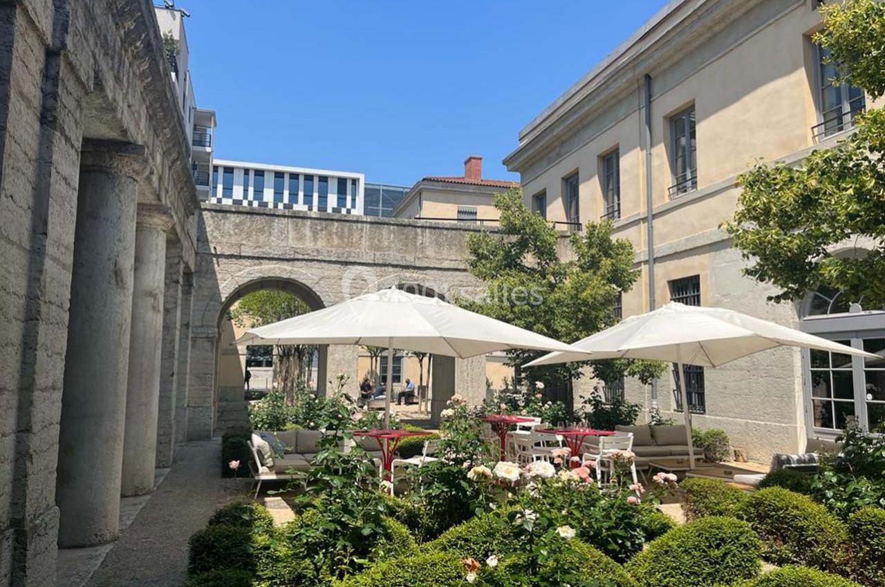 Cour intérieure avec jardin fleuri, tables et chaises sous des parasols blancs, entourée de bâtiments en pierre.