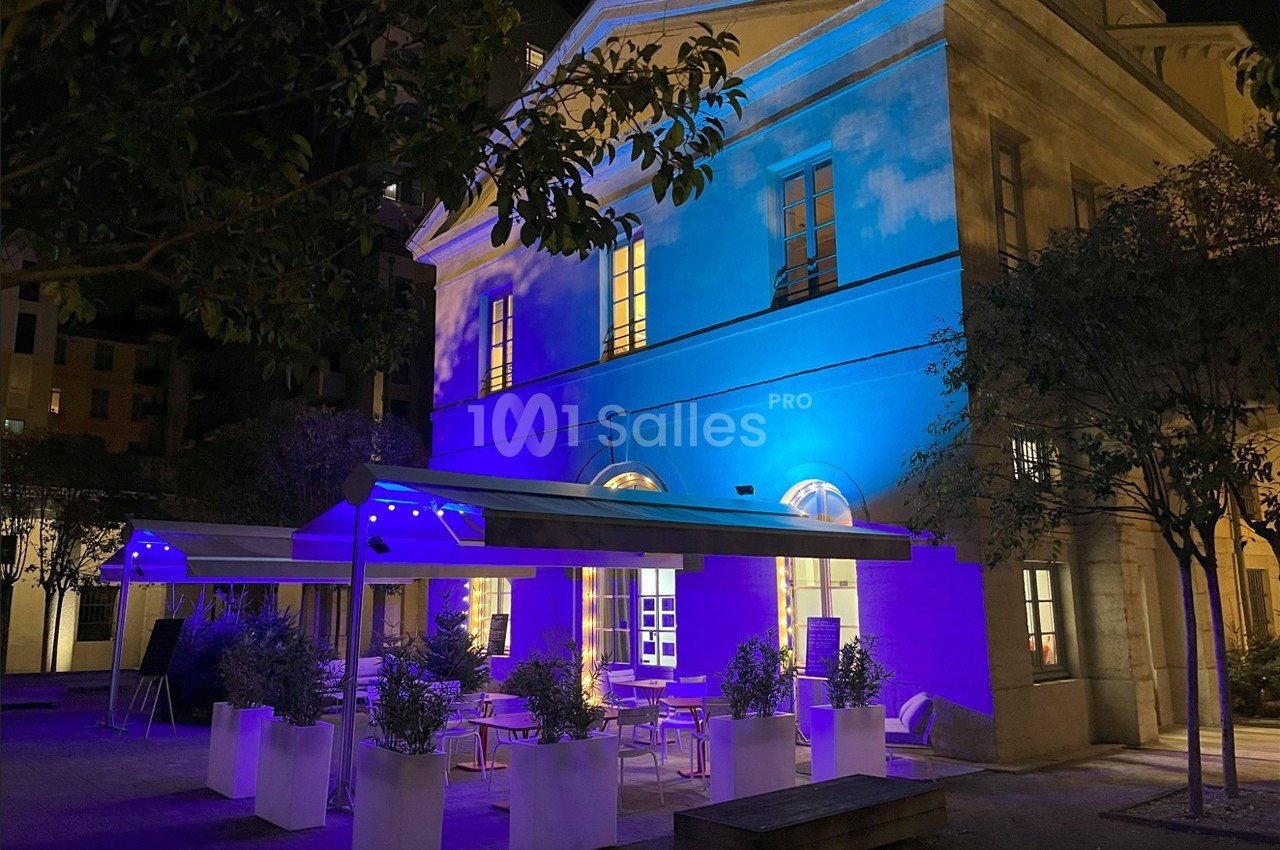 Bâtiment illuminé en bleu la nuit, avec une terrasse aménagée de tables, chaises et plantes en pots blancs.