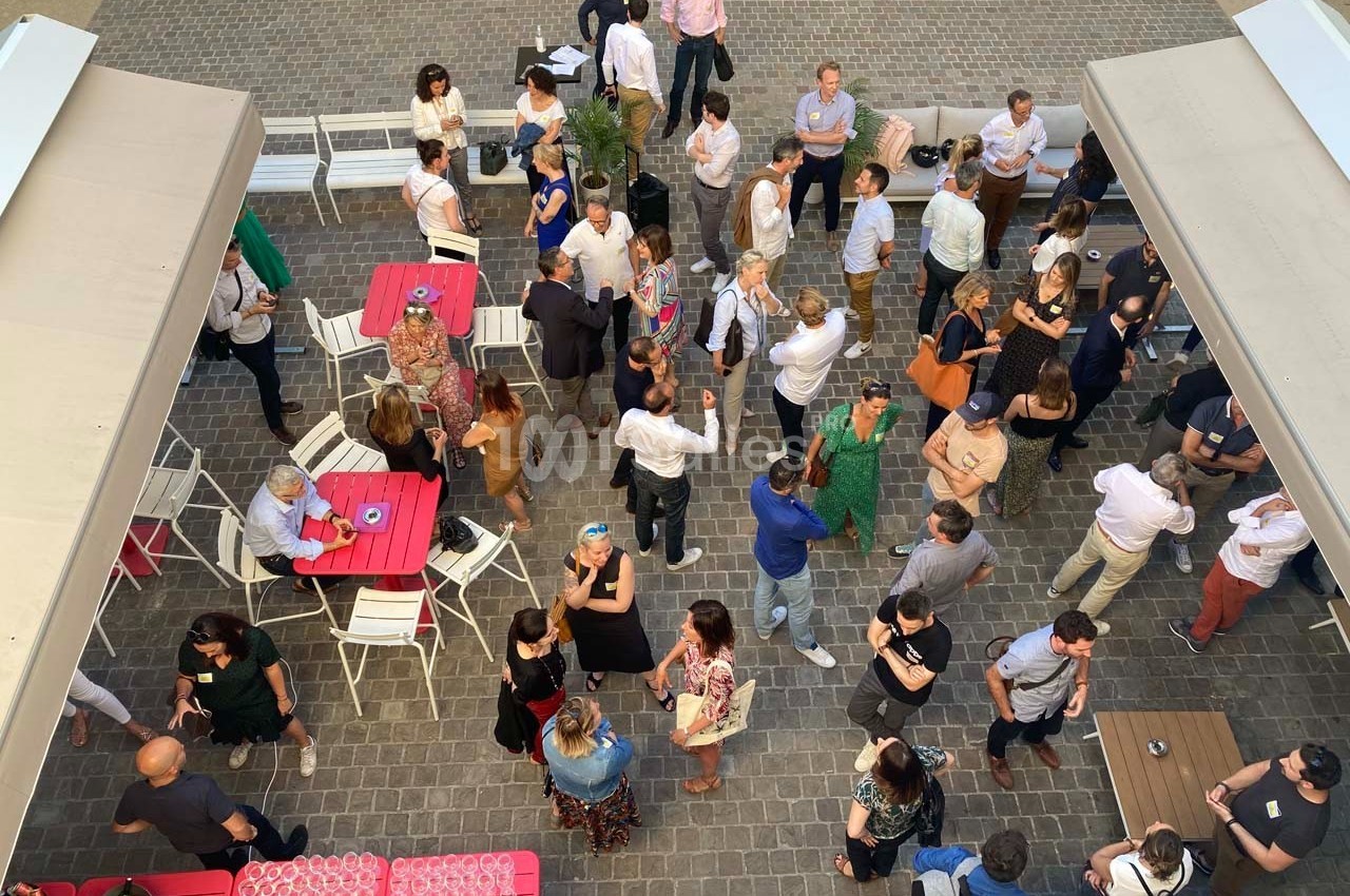 Vue aérienne d'un rassemblement en extérieur, des personnes discutent autour de tables sur une terrasse pavée.