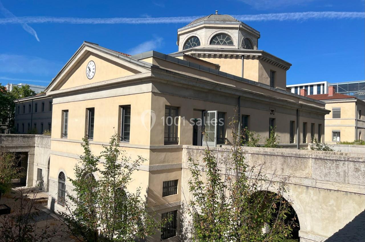 Bâtiment ancien avec façade beige, horloge en fronton et dôme central, entouré de végétation sous un ciel bleu.