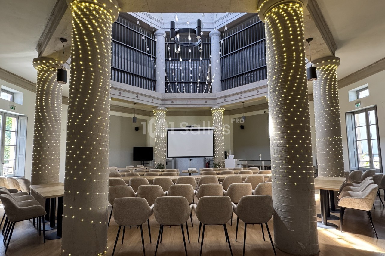 Salle de conférence lumineuse avec des colonnes décorées de guirlandes lumineuses et des rangées de chaises beige.