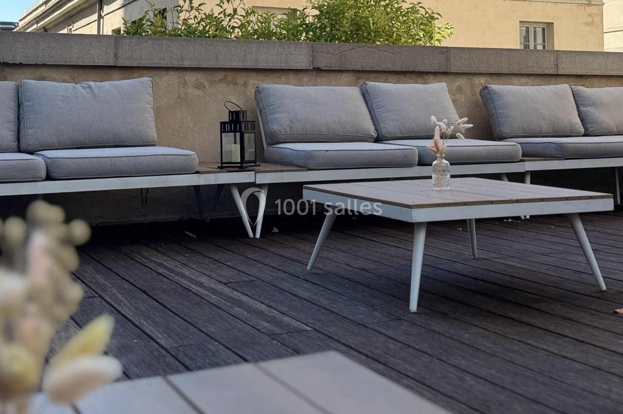 Bancs gris avec coussins et table basse en bois sur une terrasse en bois, décorée d'une lanterne et d'un vase minimaliste.