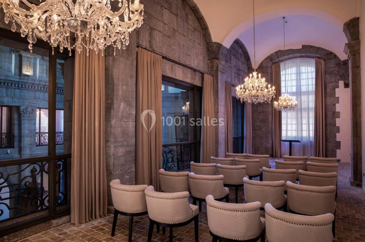 Salle avec des chaises alignées, lustres en cristal suspendus et grandes fenêtres encadrées de rideaux beiges.