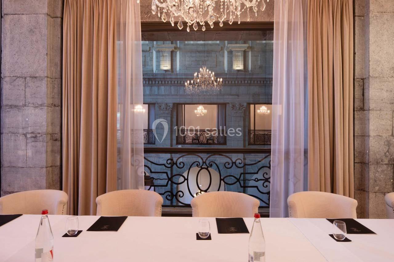 Salle élégante avec table blanche, chaises beiges, rideaux ouverts et vue sur un bâtiment illuminé par des lustres.