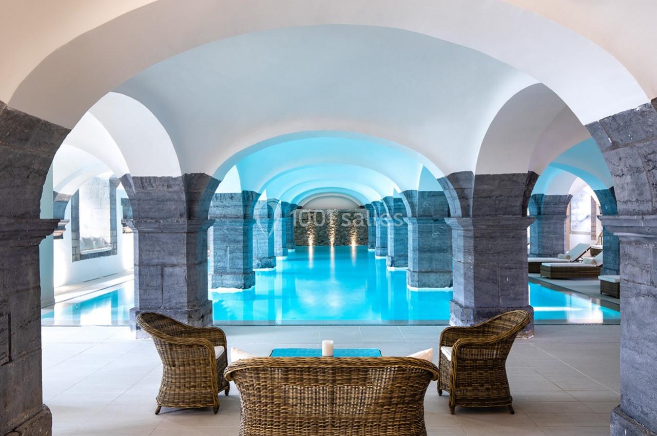 Piscine intérieure entourée d'arches en pierre, avec des fauteuils en osier au premier plan.