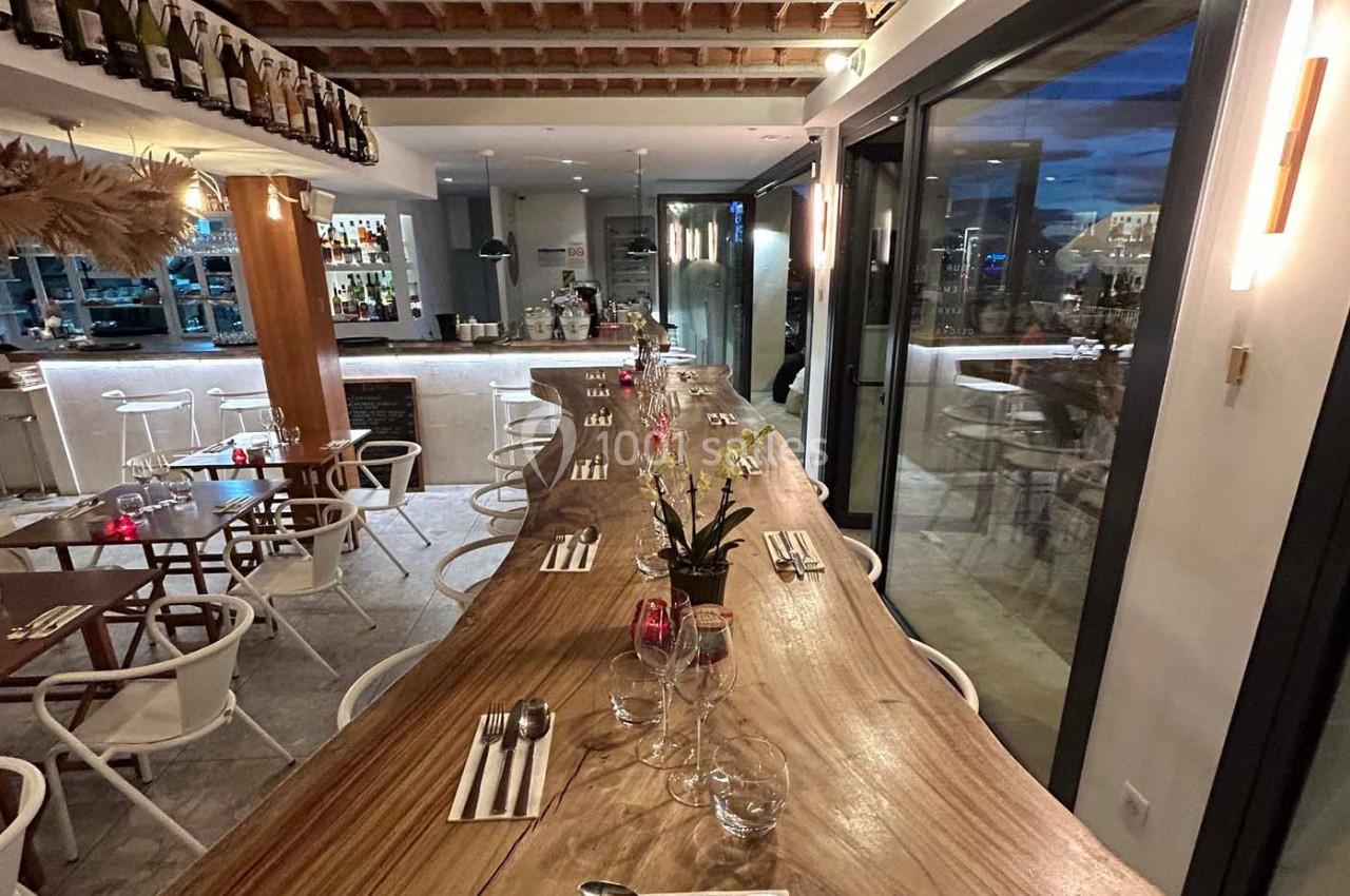 Salle de restaurant moderne avec une grande table en bois, des chaises blanches et un bar éclairé en arrière-plan.