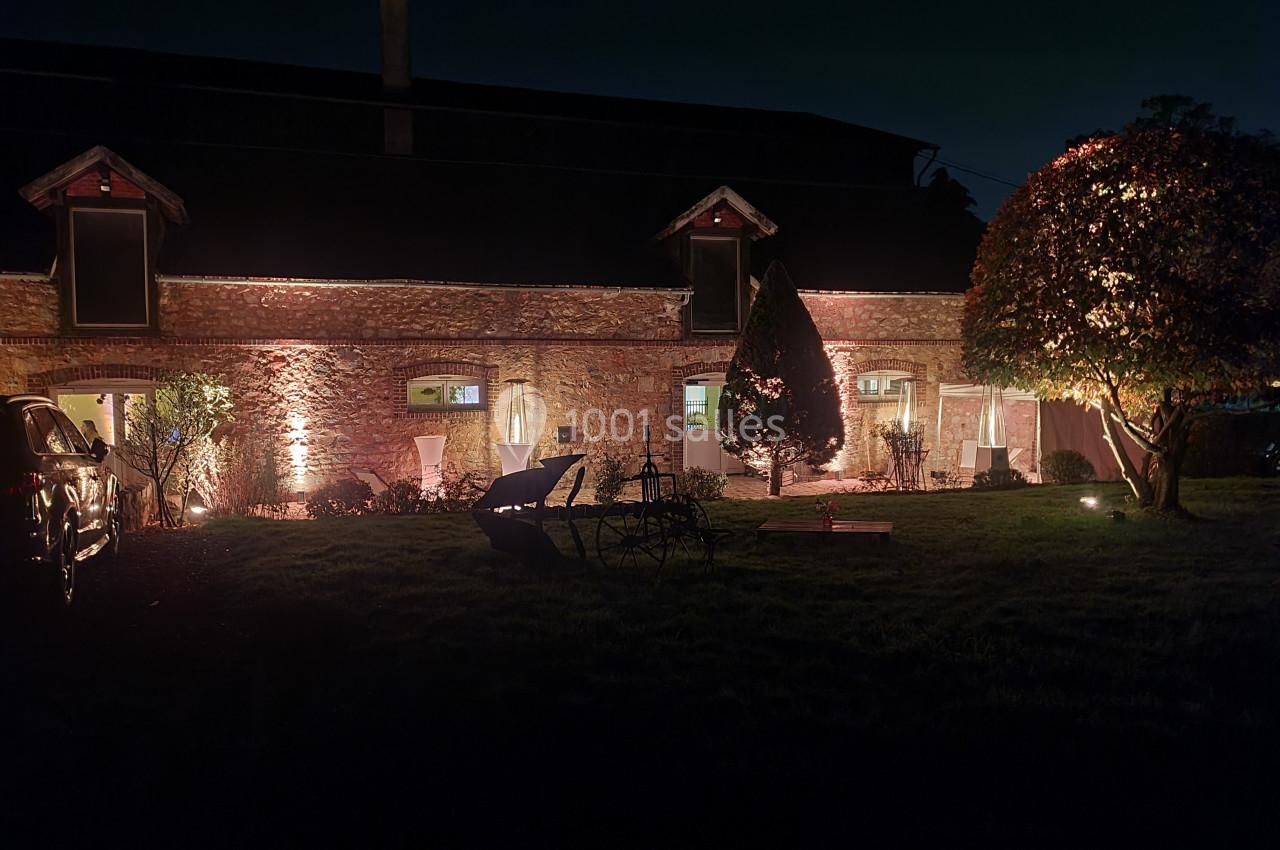Façade en pierre d'une maison éclairée la nuit, entourée d'arbres, de mobilier de jardin et d'une pelouse.