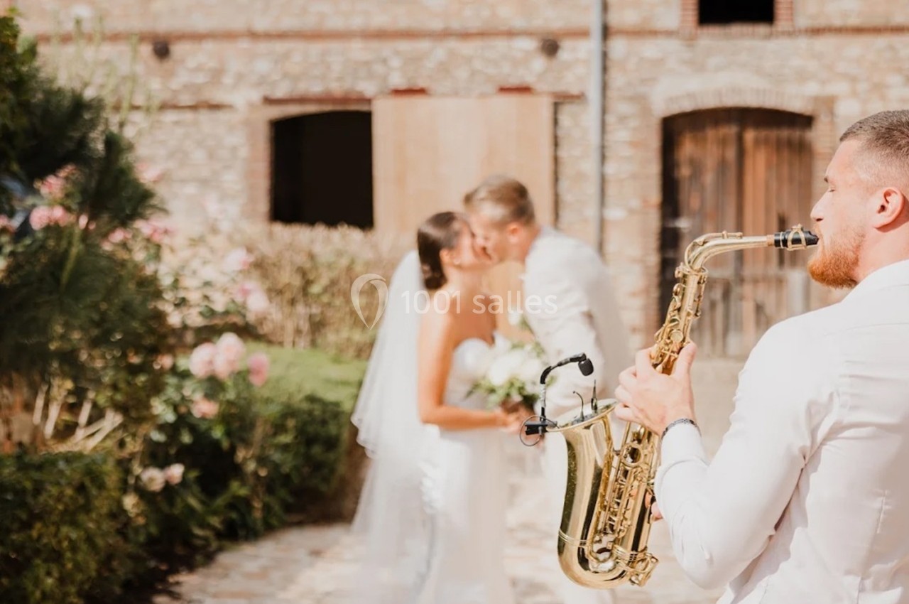 Un saxophoniste joue en extérieur tandis qu'un couple de mariés s'embrasse devant un bâtiment en pierre.