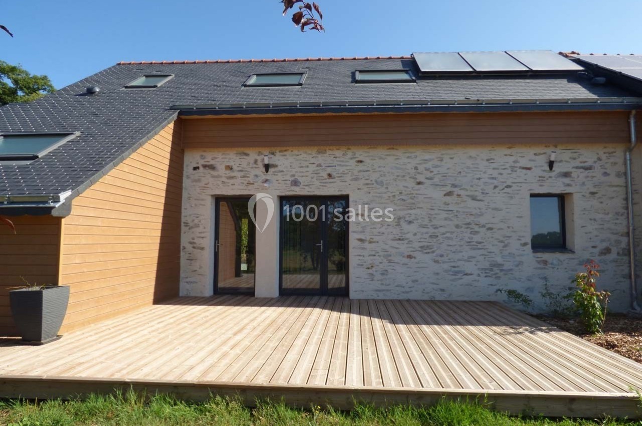 Façade d'une maison rénovée avec un mélange de bois et de pierre, terrasse en bois et panneaux solaires sur le toit.