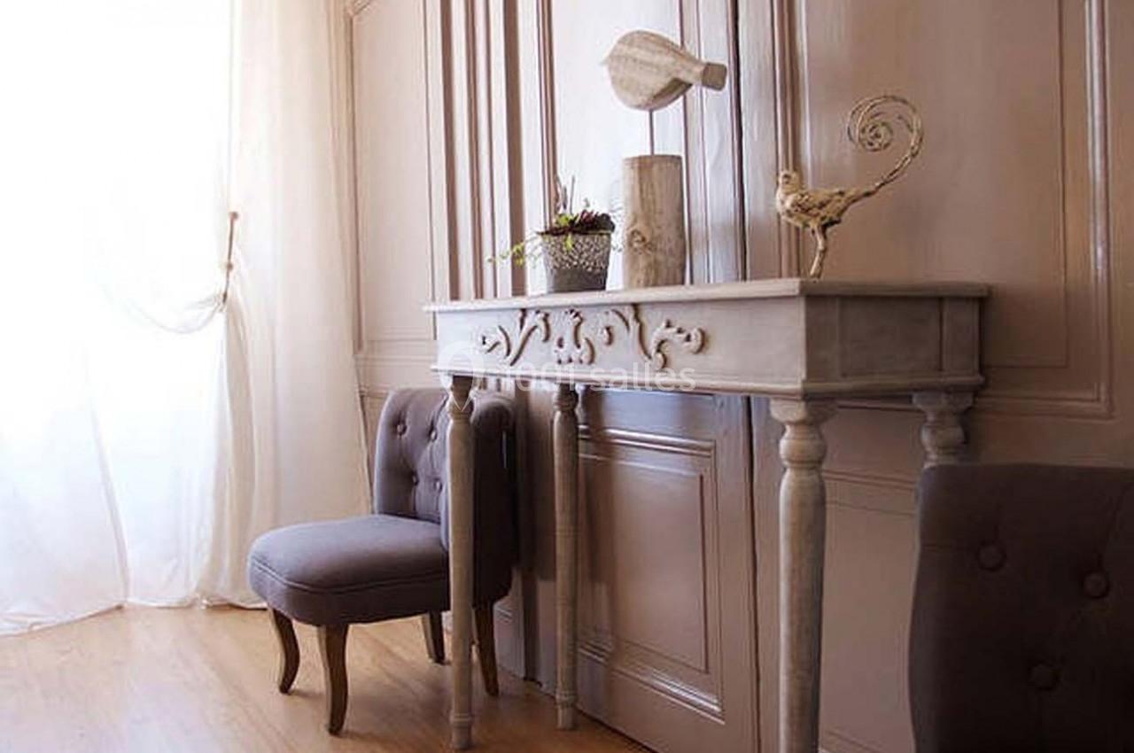 Console en bois sculpté avec décorations, chaise capitonnée grise et rideaux blancs dans une pièce lumineuse.