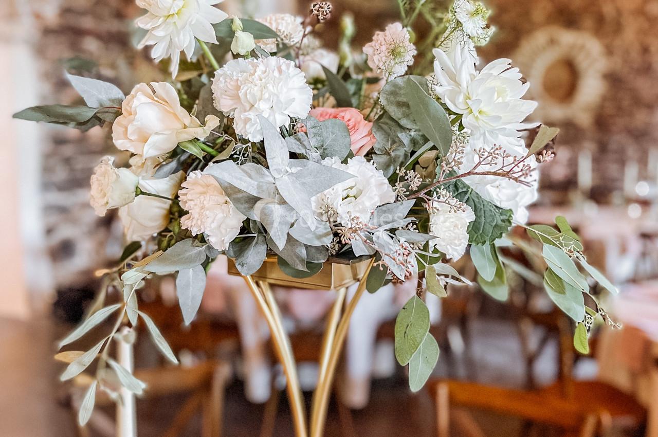 Centre de table floral avec des fleurs blanches et feuillage, posé sur un support doré dans une salle décorée de guirlandes…