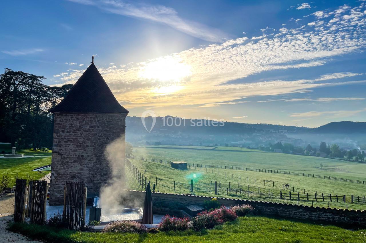 Paysage champêtre au lever du soleil avec une tour en pierre, des champs verdoyants et une légère brume.