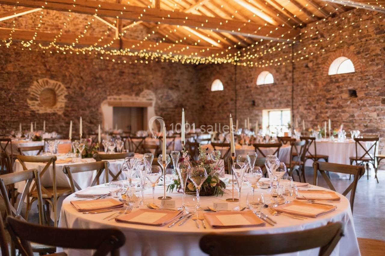 Salle en pierre avec tables rondes décorées pour un événement, éclairée par des guirlandes lumineuses suspendues.