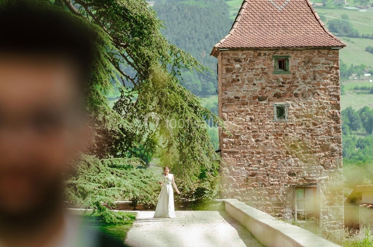 Une mariée en robe blanche se tient près d'une tour en pierre dans un paysage verdoyant, flou au premier plan.