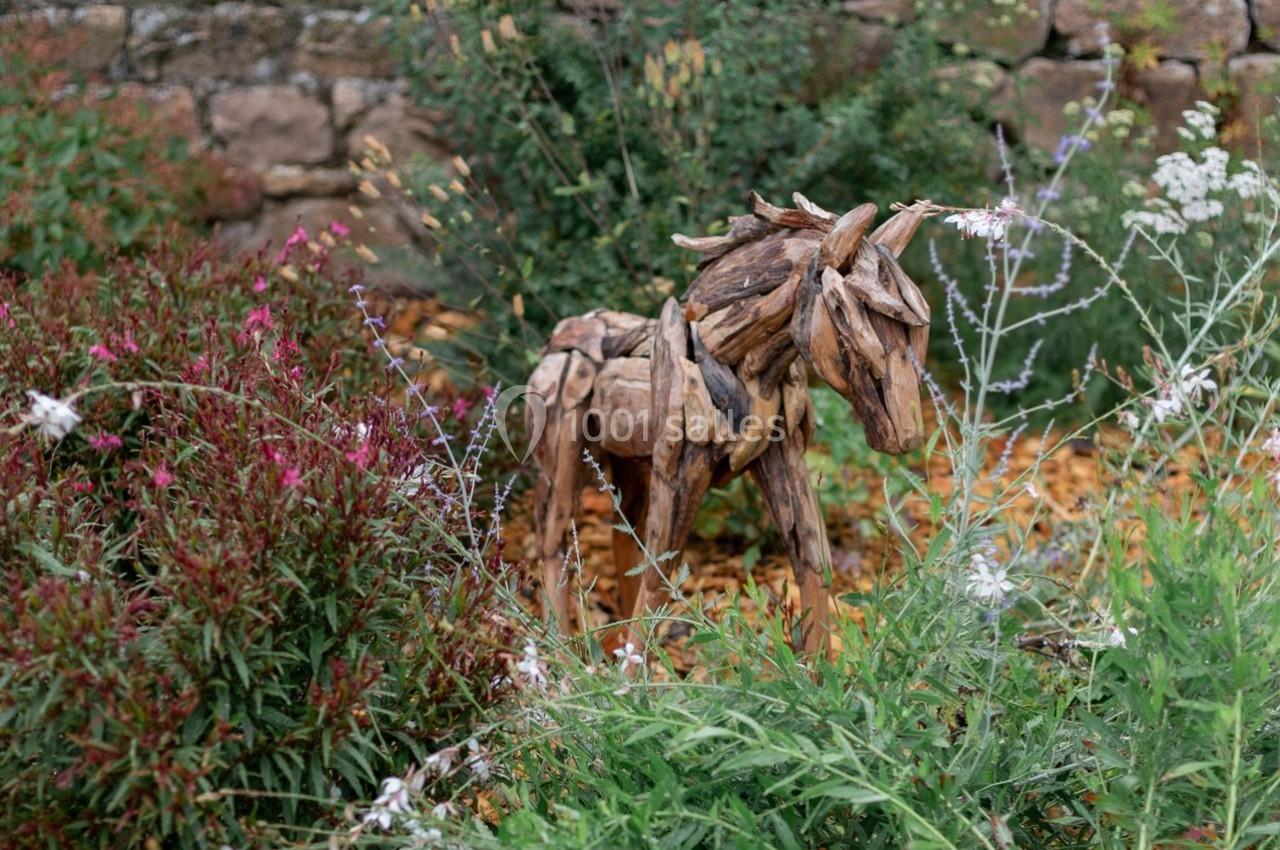 Sculpture de cheval en bois flotté entourée de plantes et de fleurs dans un jardin avec un mur de pierre en arrière-plan.