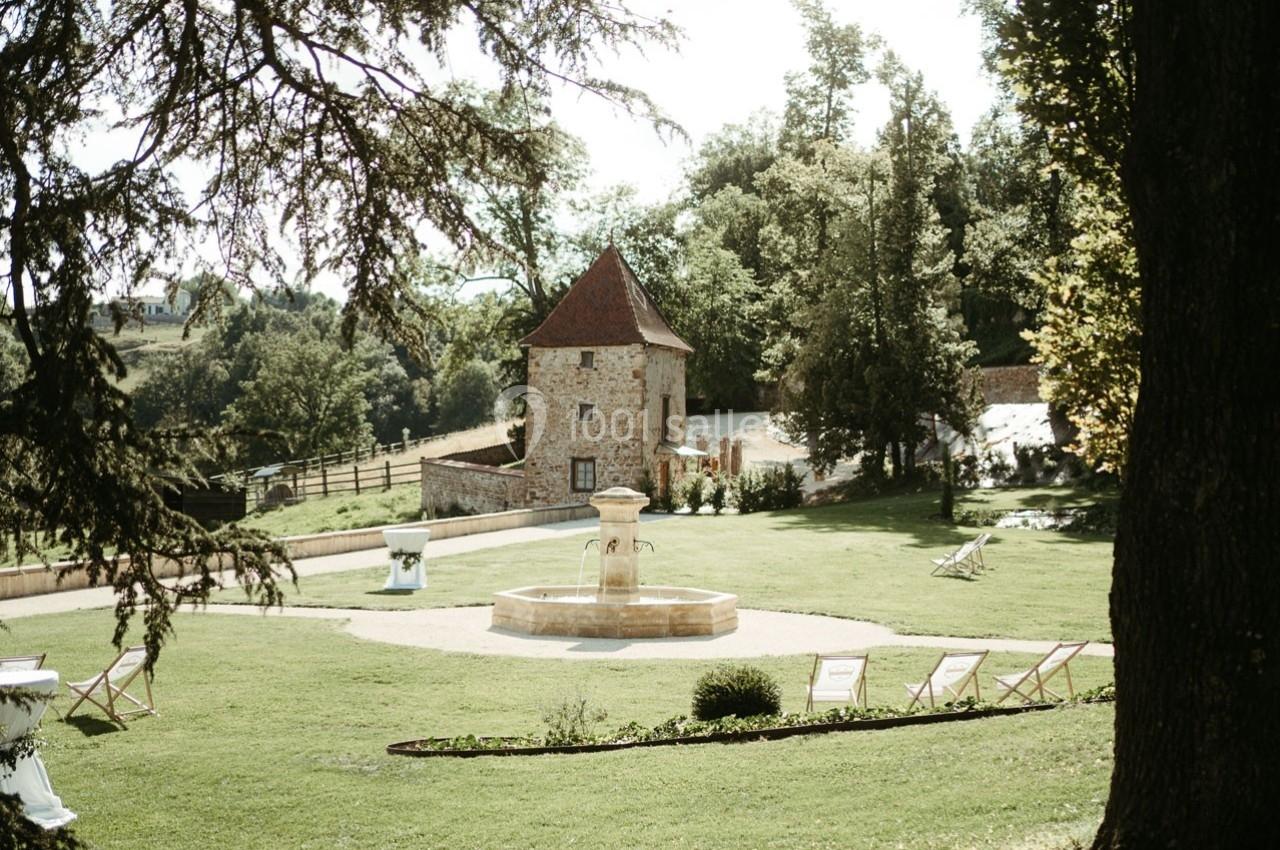 Jardin verdoyant avec une fontaine centrale, entouré de chaises longues et d'un bâtiment en pierre au fond.