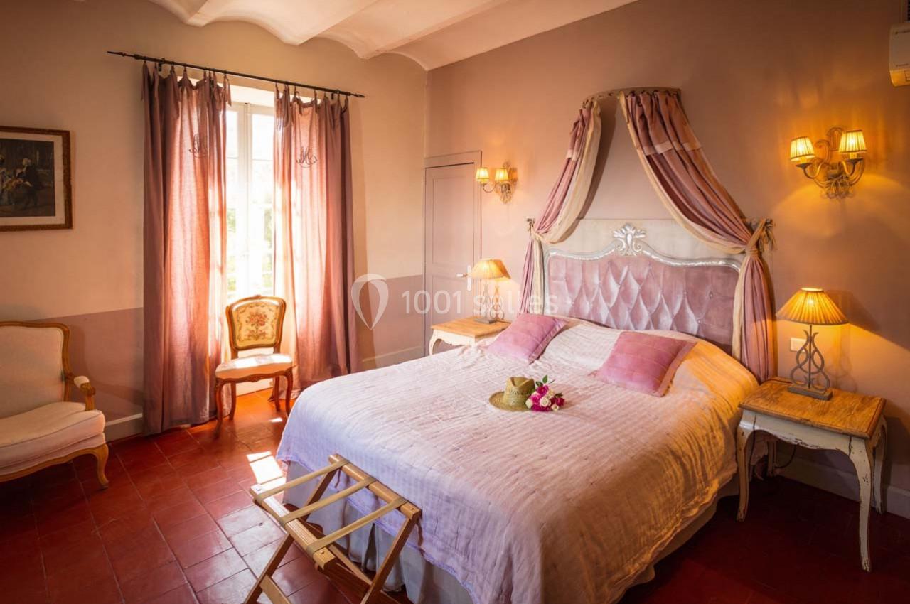 Chambre lumineuse avec lit double, baldaquin rose, meubles classiques, et lumière naturelle entrant par une fenêtre.