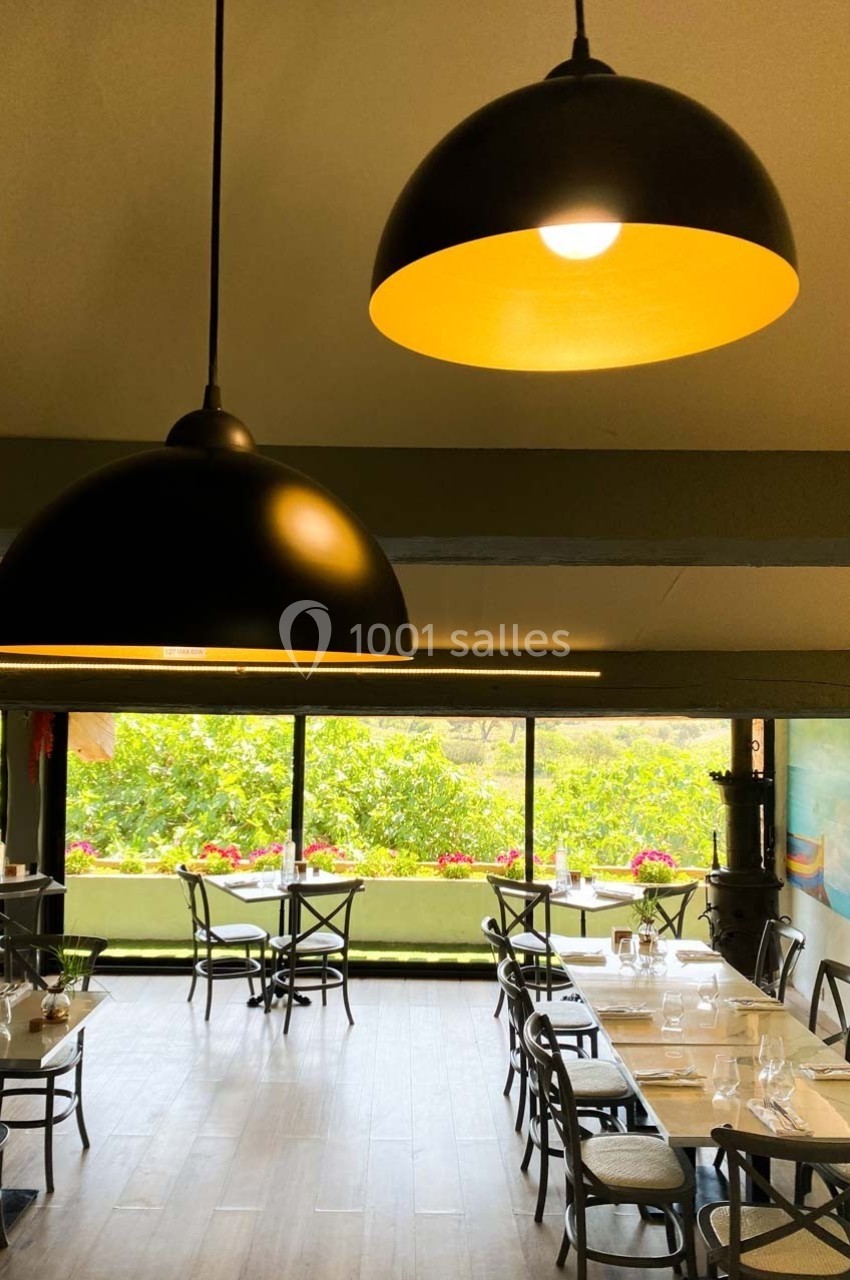 Salle de restaurant lumineuse avec grandes baies vitrées, tables dressées et suspensions noires au plafond.