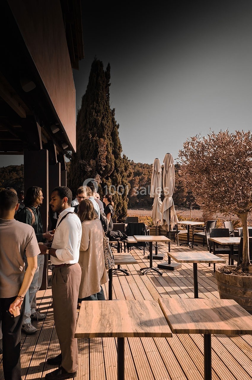 Des personnes discutent sur une terrasse en bois avec des tables, des parasols fermés et des arbres en arrière-plan.