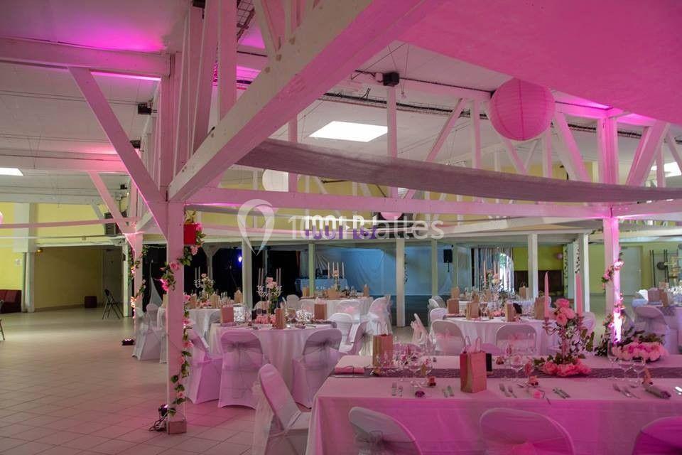 Salle de réception décorée avec des tables rondes, nappes blanches, fleurs et éclairage rose.