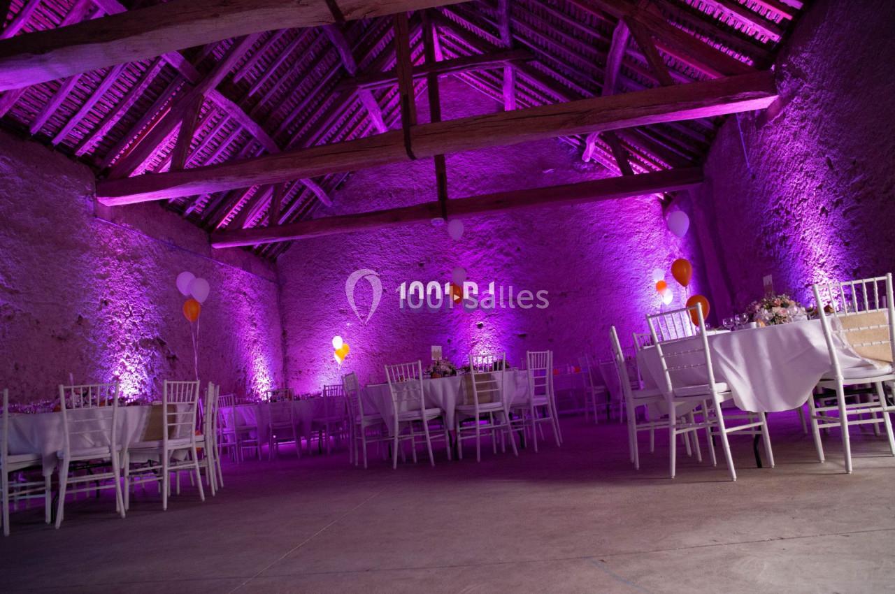 Salle de réception en pierre avec poutres apparentes, éclairée par des lumières violettes, tables et chaises blanches…
