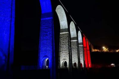 Structure en bois d'une halle éclairée par des lumières violettes, avec un toit en tuiles et des piliers en pierre.