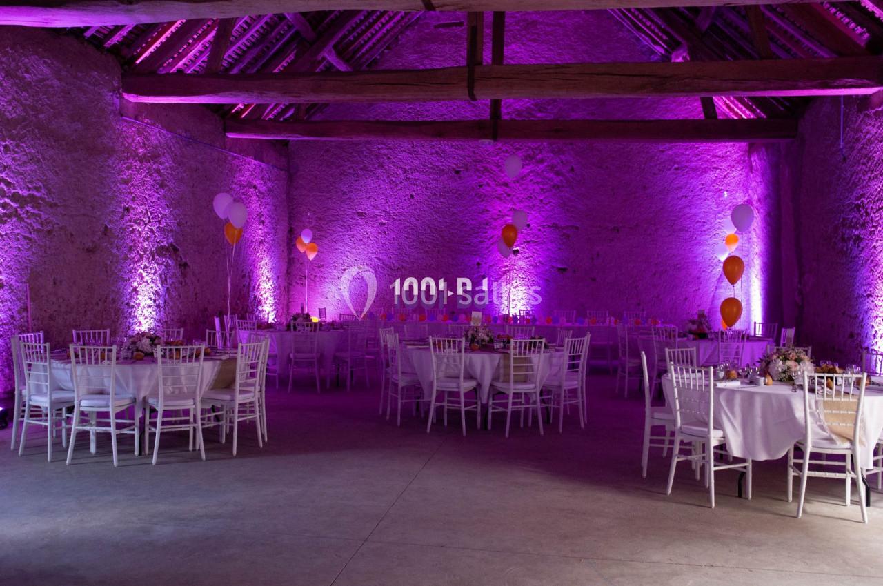 Salle de réception décorée pour un événement, avec tables rondes, chaises blanches et éclairage violet.