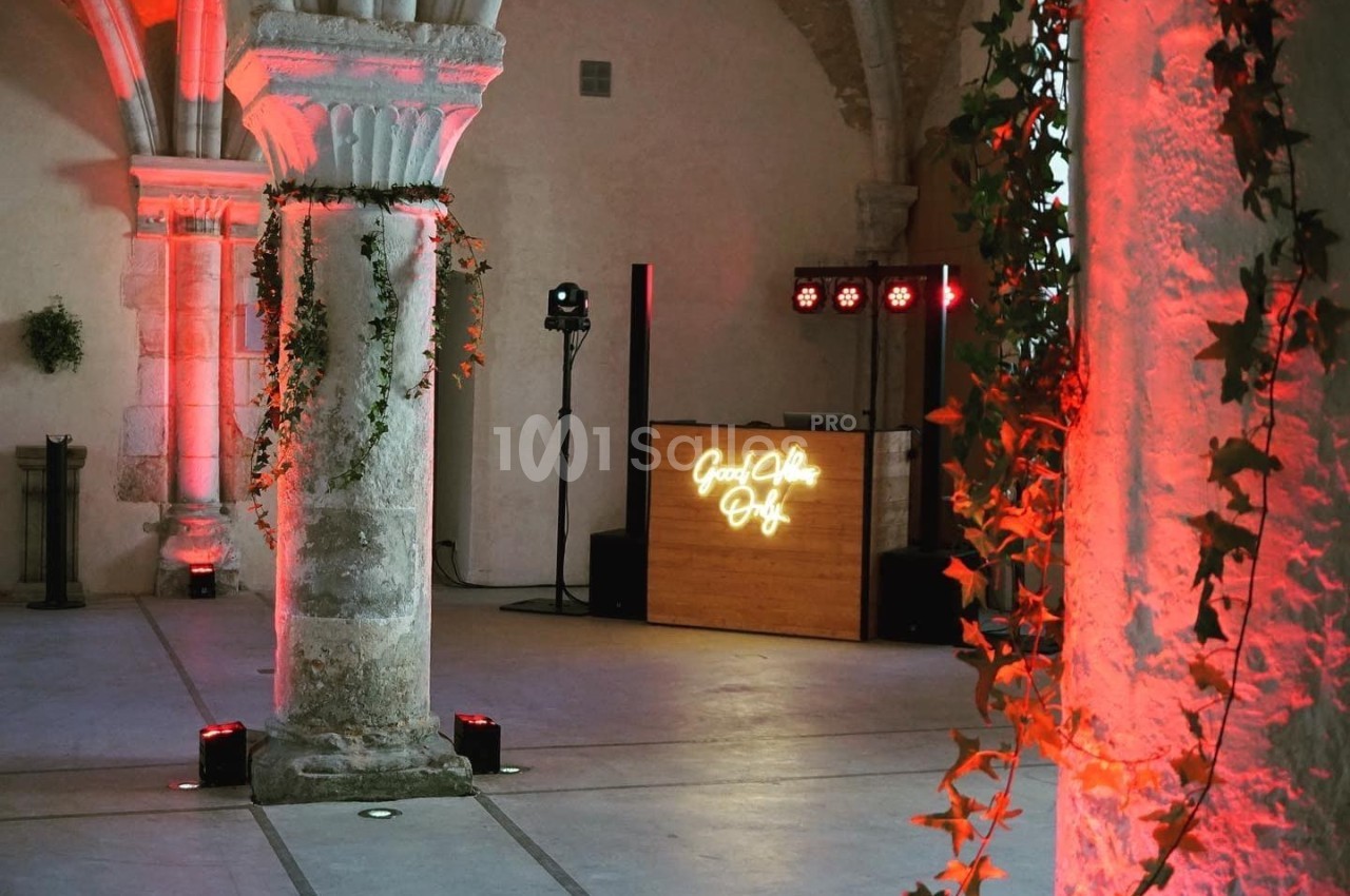 Salle voûtée avec colonnes en pierre, décorée de guirlandes végétales et éclairée par des lumières rouges et un DJ en…
