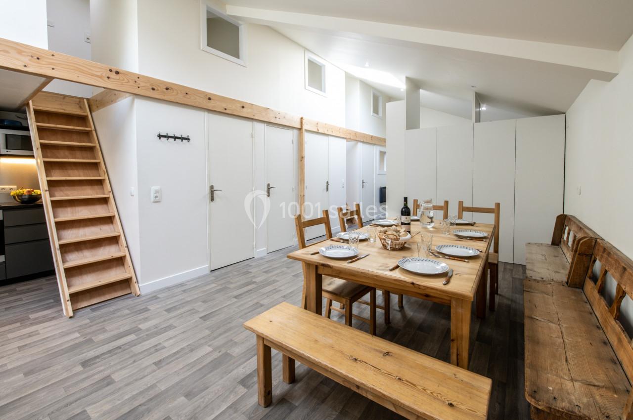 Salle à manger lumineuse avec table en bois dressée, bancs, chaises et escalier menant à une mezzanine.