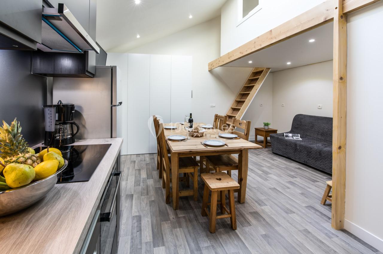 Cuisine ouverte sur une salle à manger avec table en bois, chaises, canapé gris et mezzanine en arrière-plan.
