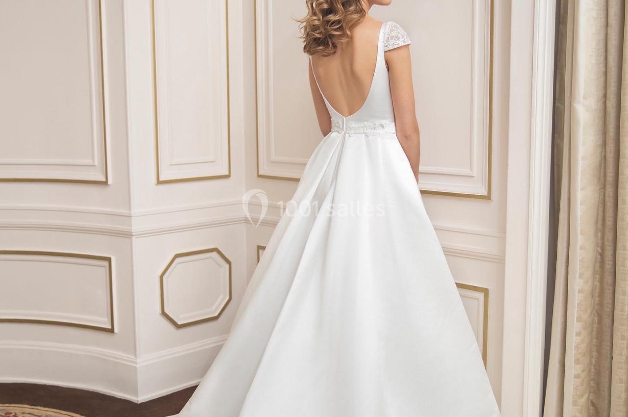 Femme portant une robe de mariée blanche à dos nu, posant dans une pièce élégante avec moulures et rideaux beiges.