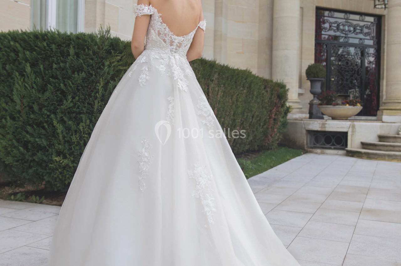 Femme en robe de mariée blanche avec dentelle, vue de dos, devant un bâtiment en pierre avec colonnes et arbustes.