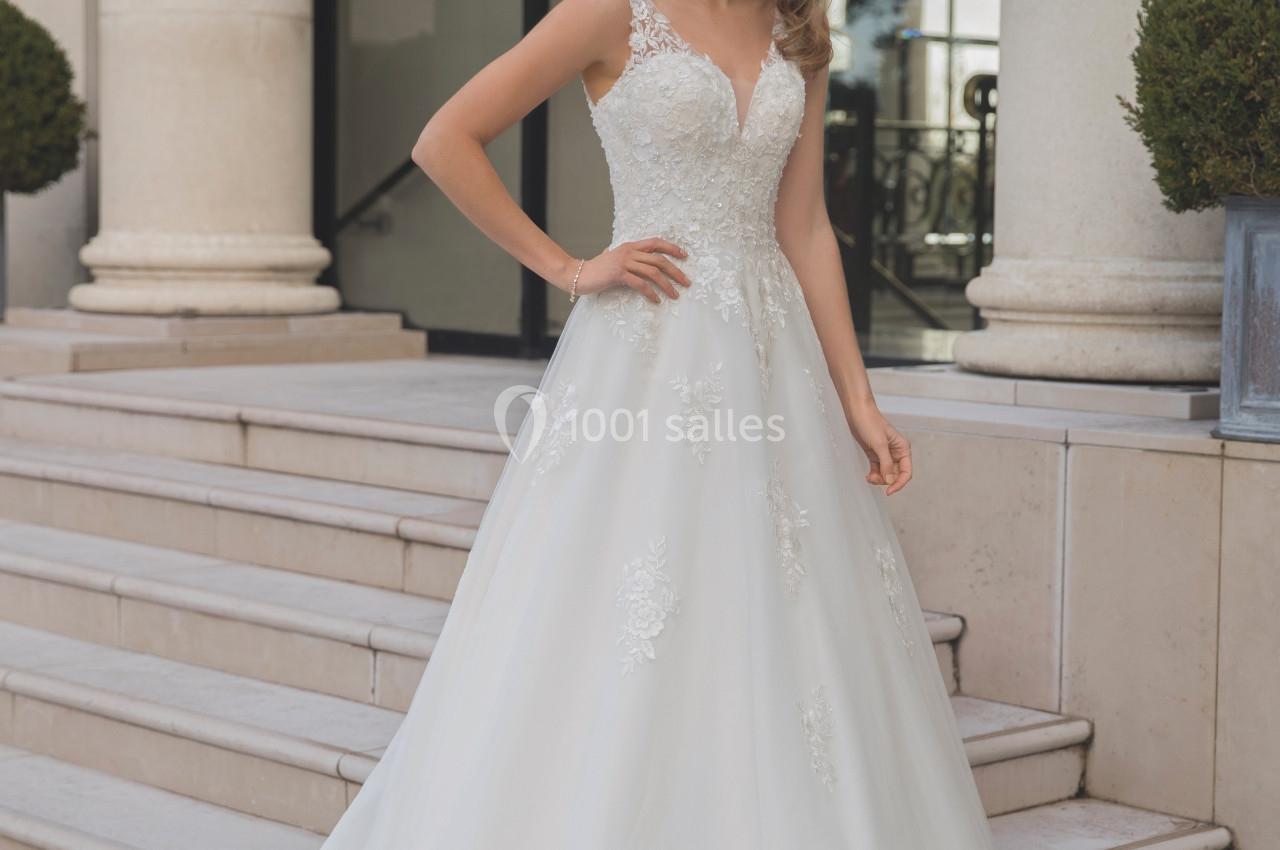 Femme portant une robe de mariée blanche avec des détails en dentelle, debout sur des marches devant un bâtiment élégant.