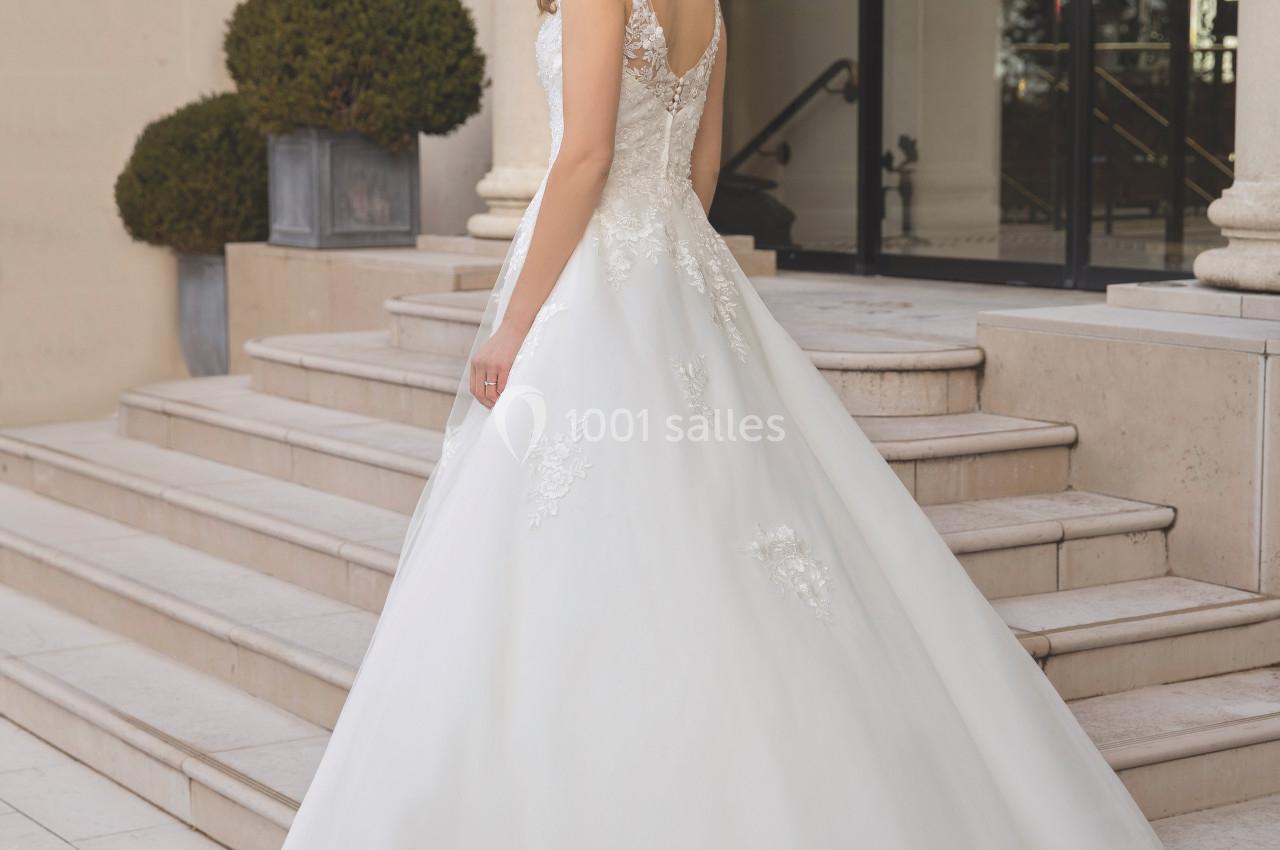 Femme portant une robe de mariée blanche avec détails en dentelle, debout sur des marches devant un bâtiment élégant.