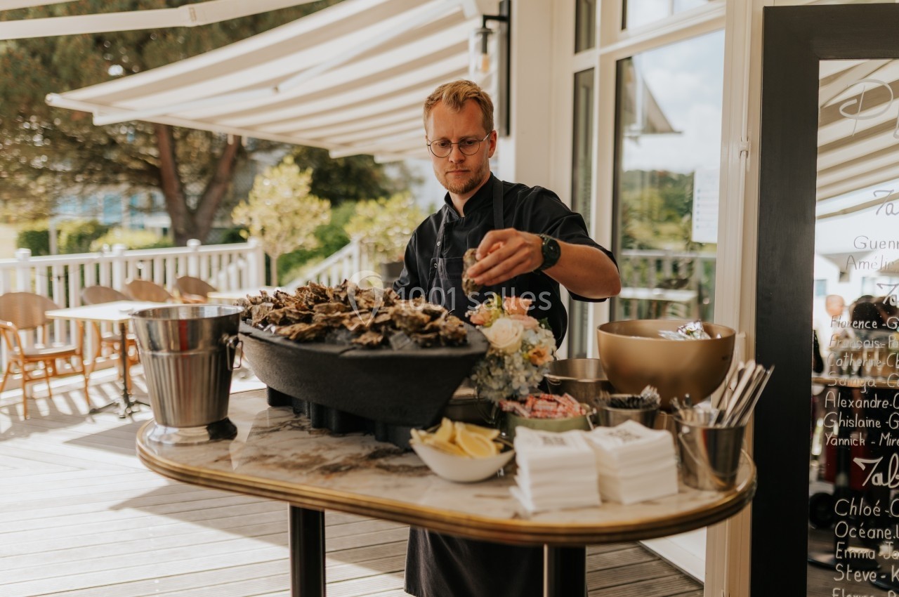 Un homme prépare un plateau d'huîtres sur une table en extérieur, entourée de fleurs et d'accessoires de service.